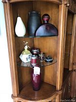 Shelf display featuring six home decor items: white and gold geometric vase with Asian script and flowers, tall black textured vase, large maroon rounded vase with handle, small ornamental vase with gold spheres, red and silver canister, black and cream floral pot with lid, and large deep red vase below.
