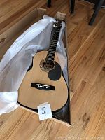 Acoustic guitar in cardboard box with white protective wrap on a wooden floor.