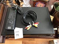 Photo of Sony DVD/CD player with remote control and RCA cables on a wooden floor surface, with auction tag number 75.