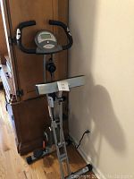 Foldable Exerpeutic therapeutic fitness bike with digital monitor on handlebar, placed next to wooden cabinet.