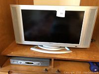Apex 27-inch widescreen flat-screen TV with silver frame and speakers on both sides, standing on a curved base. TV is shown on a wooden shelf with a remote design visible at the bottom part of the photo.
