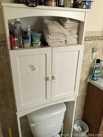 White over-toilet bathroom vanity cabinet in bathroom setting with various toiletries and towels placed on shelves.