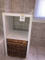 White open back shelf system with one shelf and a brown wicker basket placed in the lower compartment. Unit positioned in tiled bathroom corner below window.