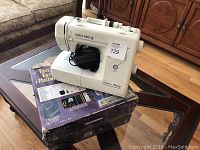 Front and top view of EURO-PRO Fast & Easy sewing machine sitting on its original box on a glass coffee table with beige patterned furniture in the background. Power cord is present.
