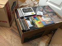 View of an open cardboard box filled with assorted music CDs in jewel cases, showing various album covers and artwork.
