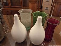 Five assorted vases on table: two white ceramic bud vases front, frosted green glass vase and green ribbed ceramic vase rear, ruby red glass vase right