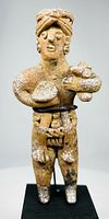 Front view of terracotta standing figure showing facial features, headdress, rounded objects held in arms, belt adornments, base