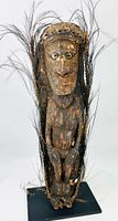 Front view of carved ancestor figure showing shell eyes, feather adornments, pigment traces, and metal display base