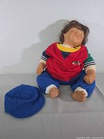 Doll seated wearing full outfit with blue cap placed beside