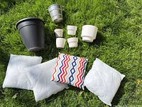 Group shot of all items on grass: black planter, metal bucket, five white pots, three pillow inserts, one chevron pillow
