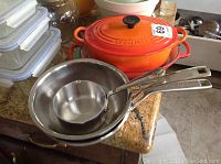 Stainless steel frying pan and saucepan nested beside orange Le Creuset cookware on countertop