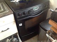 Front and side view of a black JENNAIR electric stove next to kitchen counter and trash bin, showing oven door and control panel with two rotary knobs and digital display.