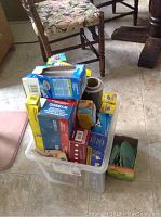 Plastic bin filled with multiple boxes and rolls of food storage supplies on kitchen floor near chair and table.