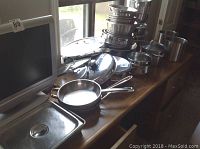Wide view showing multiple stainless steel pots, pans and lids on a wooden desk by window.