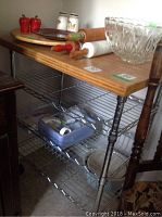 Side view of the stainless steel cart showing the wood top with various kitchen items on it and two wire shelving units with kitchenware below.