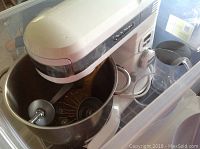 White Cuisinart stand mixer inside large plastic bin with Stainless steel bowl and three mixer attachments visible