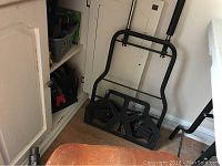 Metal dolly with two wheels and foldable handle resting against wall, adjacent shelf visible.