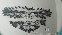 Photo showing two metal wall decor pieces mounted on wall, designed with multiple leaf elements in blue, brown, and gray tones. Larger piece on top, smaller piece below.