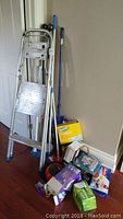 Photo showing aluminum multi-step ladder leaning against wall, Swiffer duster refill yellow box, dust mop, basket, and cleaning supply boxes on floor.