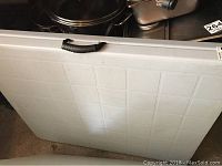 Photo showing full view of the folded white plastic folding table with integrated black handle and grid pattern surface