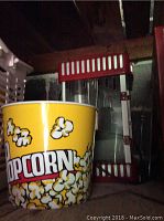 Photo showing a yellow popcorn container with popcorn graphics and the popcorn maker machine behind it.