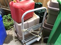 View of the metal utility cart from front with red gas can and a grey plastic bin on top, showing rust and chipped paint on the frame.