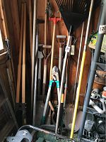 Assorted garden tools leaning against wooden wall including rakes, shovels, loppers, and a spiked wheel tool.