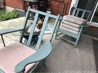 Two painted wood armchairs with pink seat cushions placed beside two small round glass-top metal side tables on a porch deck.