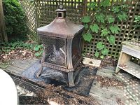 Full view of the metal chiminea in an outdoor setting on a metal pad among plants and lattice.