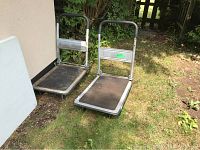 Two metal platform carts shown side by side outdoors on grass. One cart leans against a wall. Both carts have rust and wear on metal frames and rubber mats on platforms.