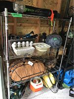 Metal shelving unit with various garden decor items and power tools including a lantern, concrete candle holders shaped like birds, a metal planter with coir liner, and toolboxes or compressors on the bottom shelf.