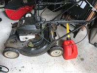 Full view of MTD lawn mower with black metal deck and white engine cover, showing push handles and cables; red plastic fuel container with yellow spout placed beside mower.