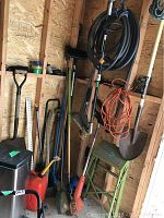 Overall view of garden tools including shovels, trimmer, hoses, extension cord, and ladder in a wooden shed