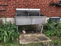 Full view of stainless steel double sink with legs installed outdoors against brick wall.