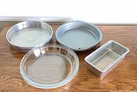 Four baking pans arranged on table: two round metal, one glass pie plate, one loaf pan