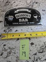 Front of black and chrome Brandon's Bar sign beside measuring tape showing 6 in width