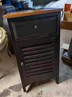 Angled front view of black nightstand with drawer, louvered door and wood top