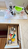 Countertop view with Pyrex measuring cups, OXO measuring cup, grater, green colander and drawer of utensils