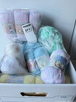 Box containing multiple sealed packs and loose skeins of pastel, blue, green, yellow, and white Baby's Delight yarn