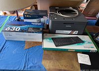 Group shot showing Brother printer, two toner cartridge boxes, Logitech K120 keyboard box, Panasonic cordless phone box, and table lamp