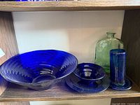 Shelf view showing large cobalt blue ribbed bowl, smaller cobalt blue plate and dish, cobalt blue cylindrical vase with base, and flat green jug bottle