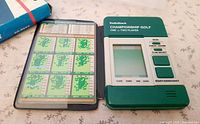 Front view of open handheld game showing controls, LCD screen, course card booklet, partial view of box