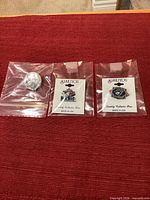 Group shot of three of the four pins in individual plastic bags on red surface