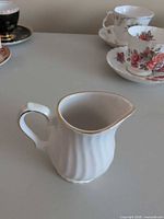 Angled front view of white ribbed creamer with gold trim, cups and saucers blurred in background