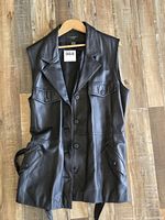 Front view of black leather vest on hanger showing buttons, lapels, pockets, waist belt