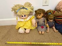Three dolls—yellow dress, grey polka-dot dress, blue romper—next to wall for scale