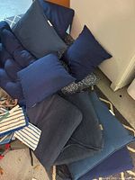 Pile of various blue cushions and pillows including tufted lounge cushion, square seat pads, solid and patterned throw pillows