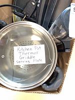 Box showing stainless pot with steamer, part of griddle and grinder visible