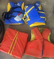 Group shot of four PFDs: two blue/yellow vests, one orange cushion, one orange/yellow vest