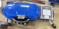 Top view of closed blue Coleman grill on wheeled cart with manual beside